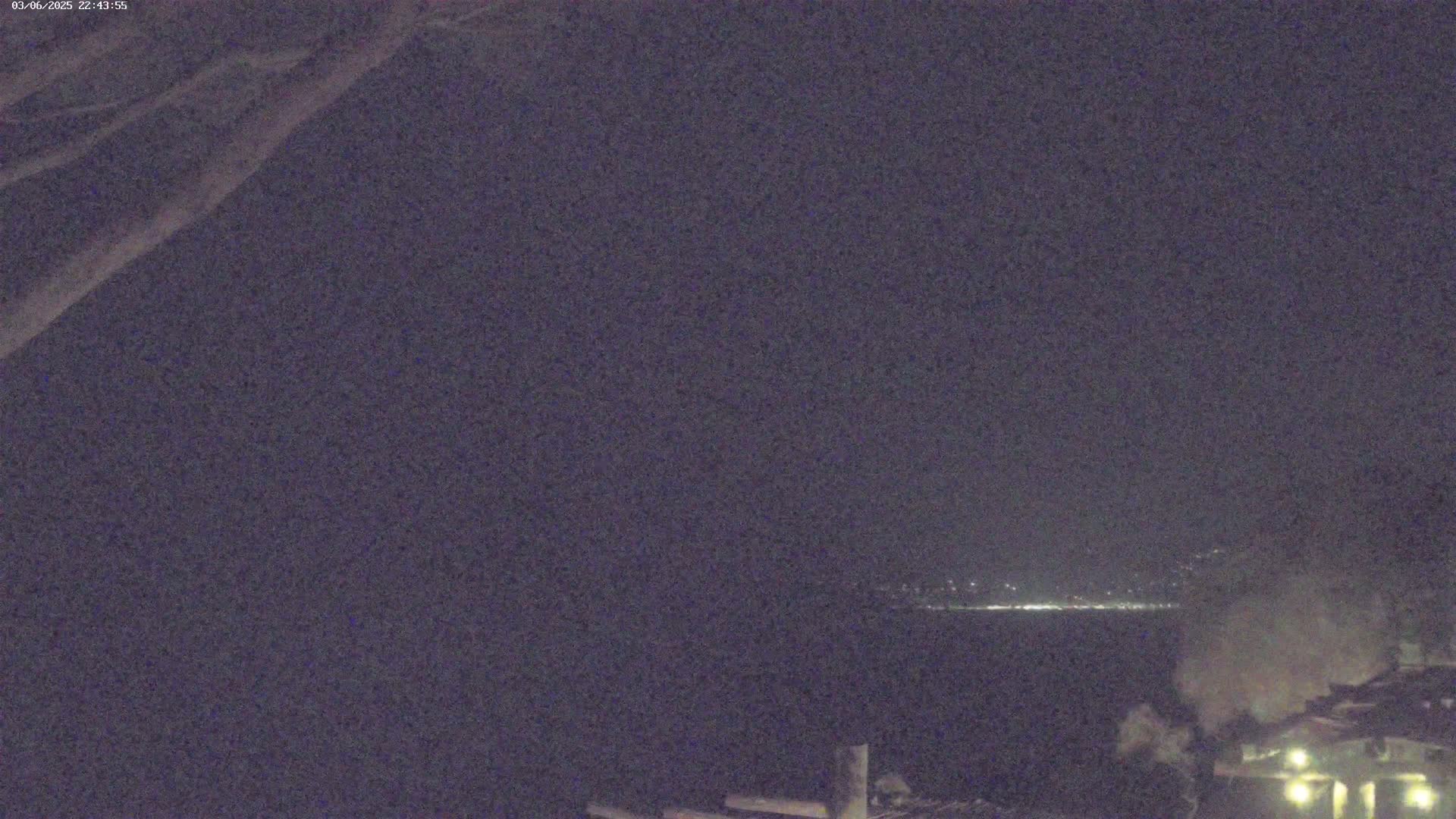 A nighttime, low-light view shows a body of water with distant lights on the far shore, some smoke or steam rising near the bottom right, and part of a building or structure visible in the lower right corner.