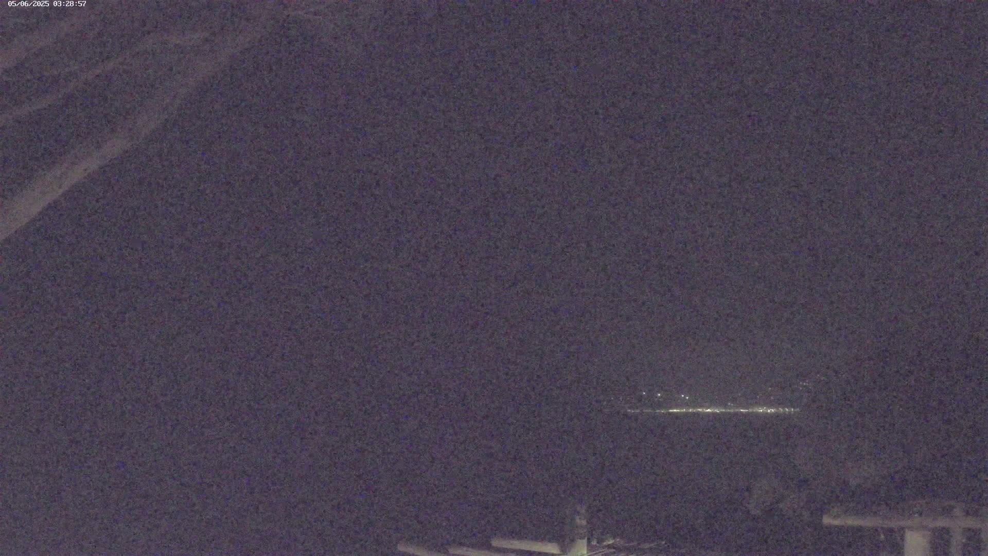 A nighttime, low-light image shows a distant line of lights across a dark body of water, with part of a dark structure visible in the bottom right corner.