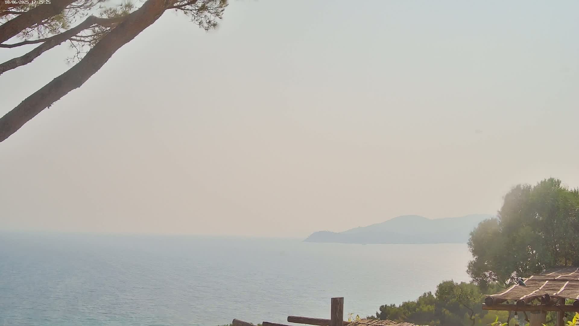 A hazy, sunny day overlooks a calm sea with distant hills and a portion of a wooden structure in the foreground.
