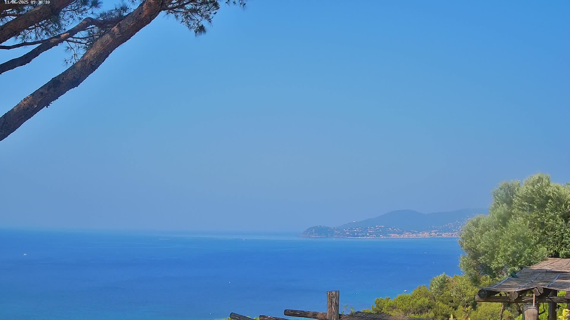 A tranquil ocean scene under a clear blue sky shows a distant coastline with low mountains, viewed from a vantage point partially obscured by tree branches and a rustic wooden structure.
