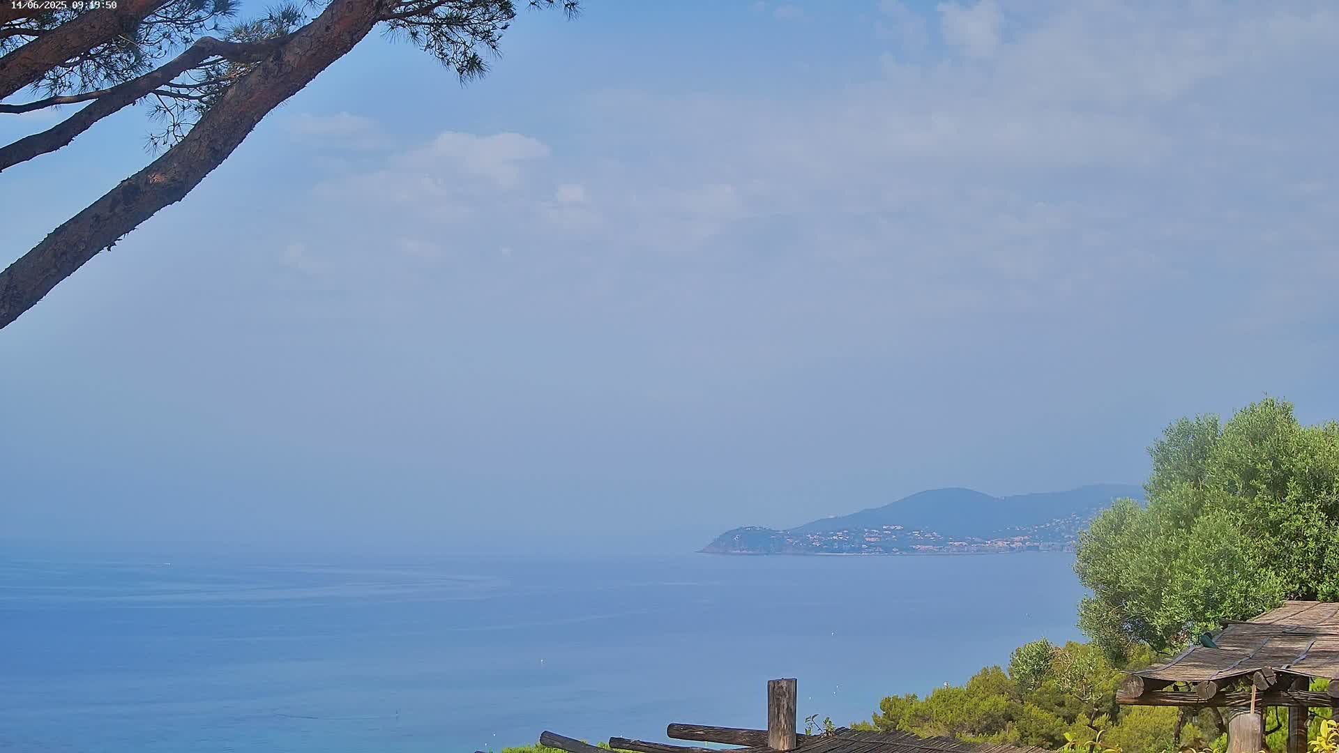 A partly cloudy sky overlooks a calm blue sea with a distant landmass, partially obscured by a tree branch in the foreground and a wooden structure to the right.