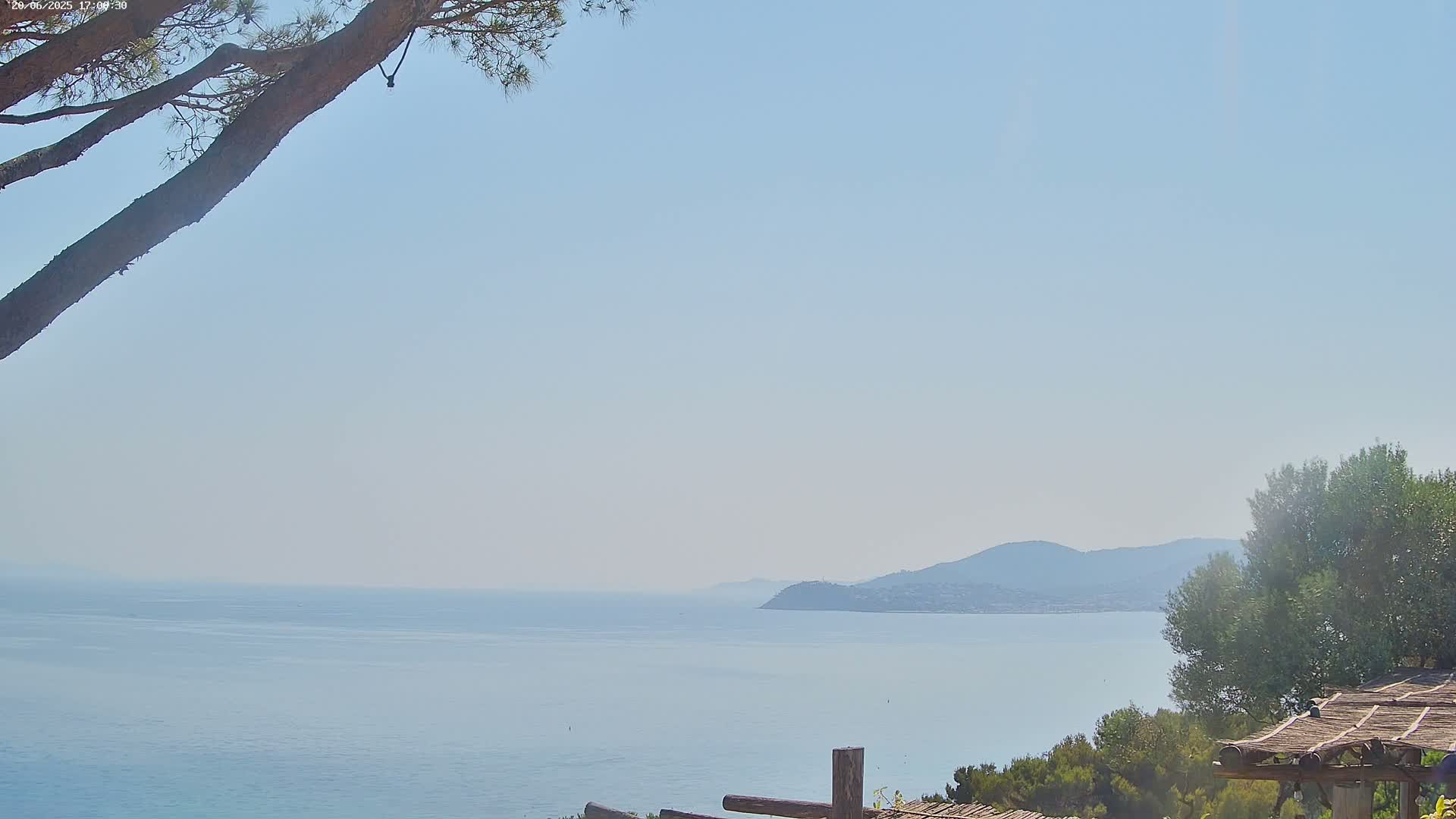 A calm, hazy ocean stretches to a distant mountain range under a clear, light-blue sky, partially obscured by tree branches and vegetation in the foreground.