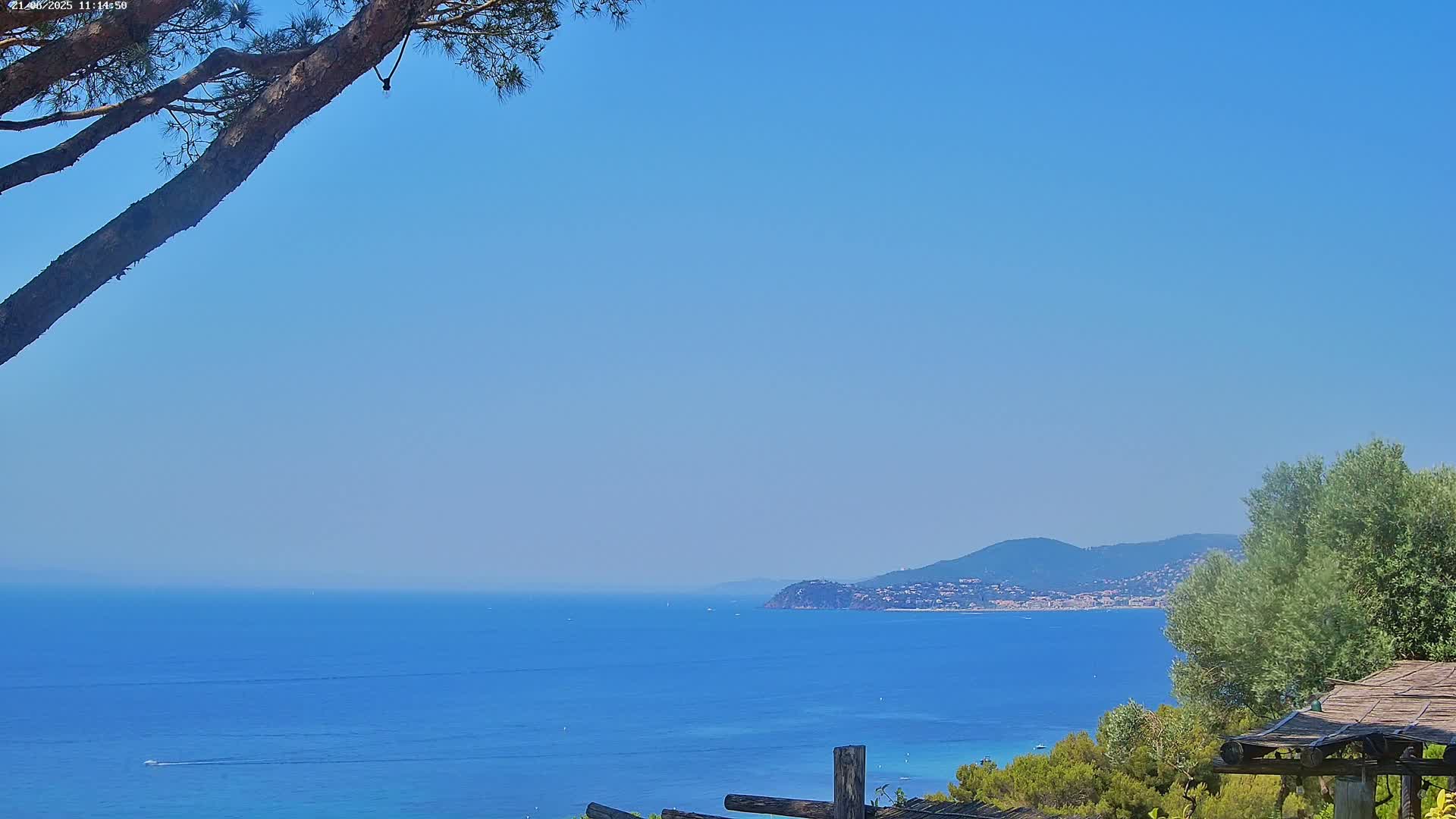 A tranquil coastal scene on a clear, sunny day shows a calm blue sea extending to a distant mountain range partially obscured by vegetation in the foreground.