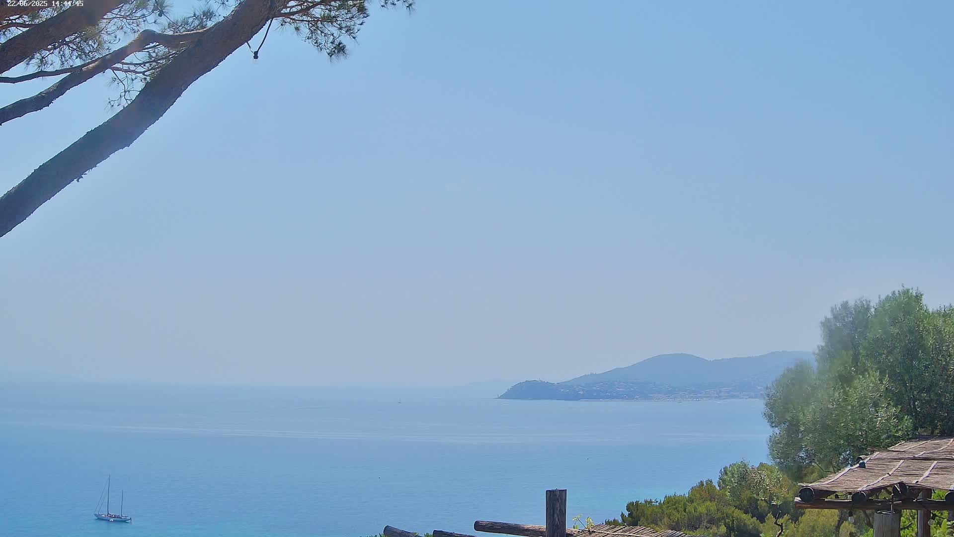 A sailboat is visible on a calm, light blue sea, with a distant mountain range under a clear sky, partially obscured by tree branches in the foreground.