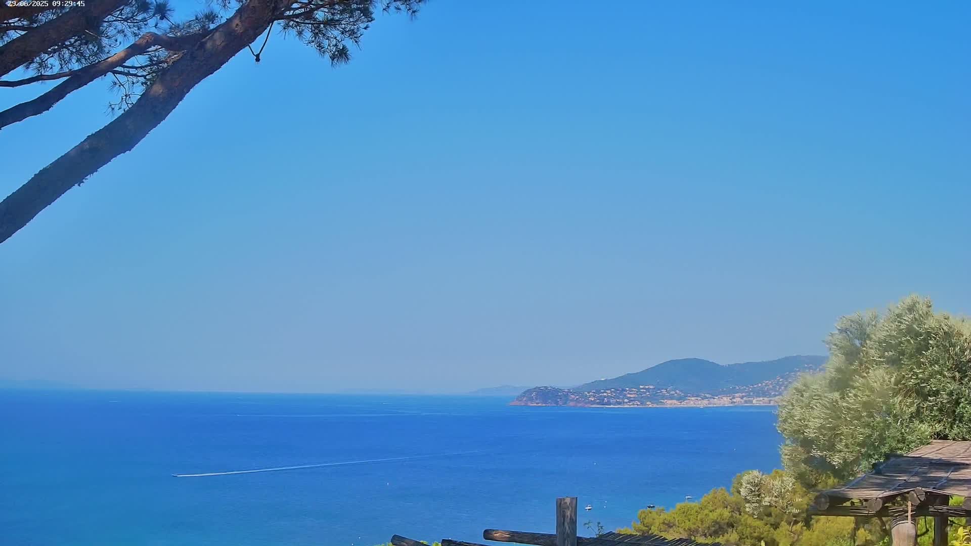 A tranquil coastal scene on a clear, sunny day shows a calm blue sea extending to a distant mountainous shoreline, partially obscured by a pine tree branch in the foreground and some shrubbery near a wooden pergola.