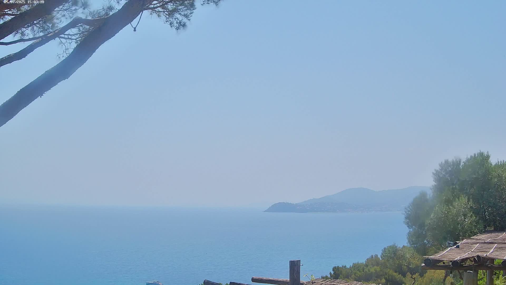 A hazy, sunny day reveals a tranquil blue sea stretching to a distant, indistinct coastline under a pale sky, partially obscured by tree branches in the foreground.