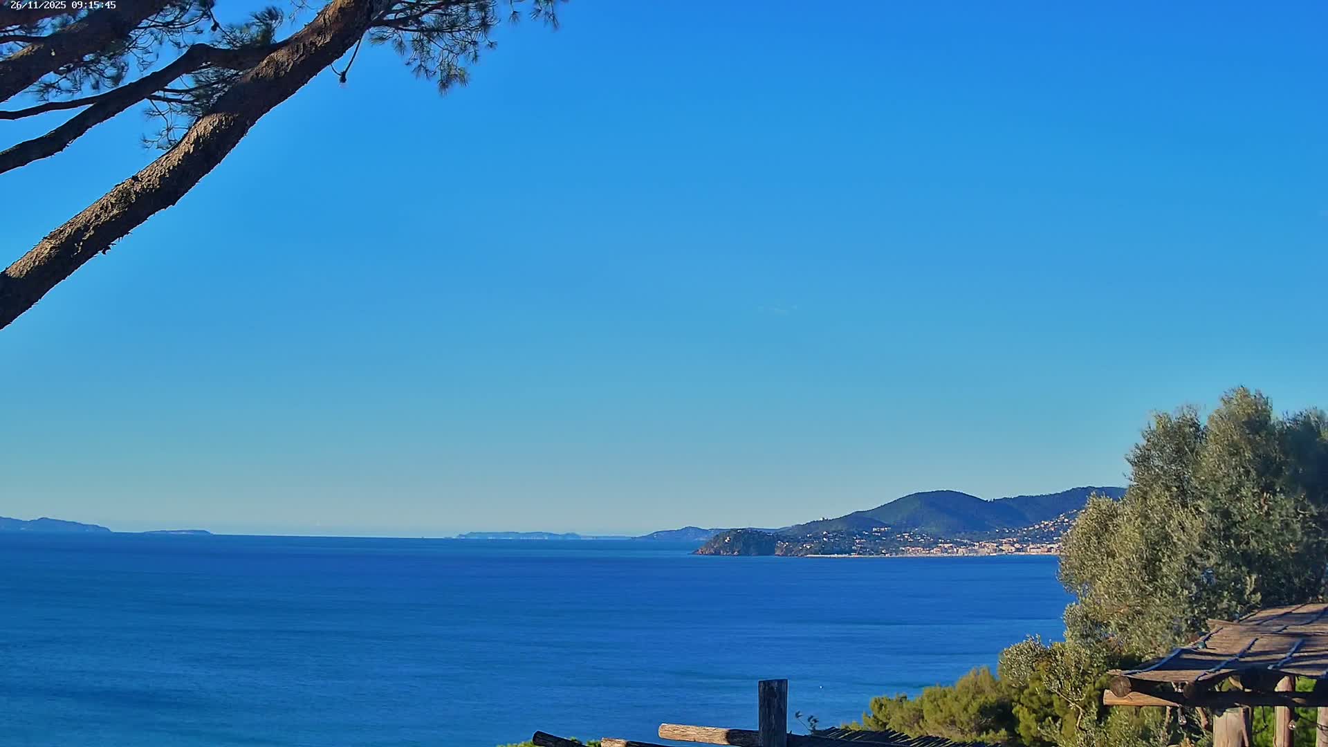 A serene coastal view features a calm, deep blue sea under a clear, bright blue sky, with distant landmasses and a town nestled along the far shore, framed by parts of trees and rustic wooden elements in the foreground.