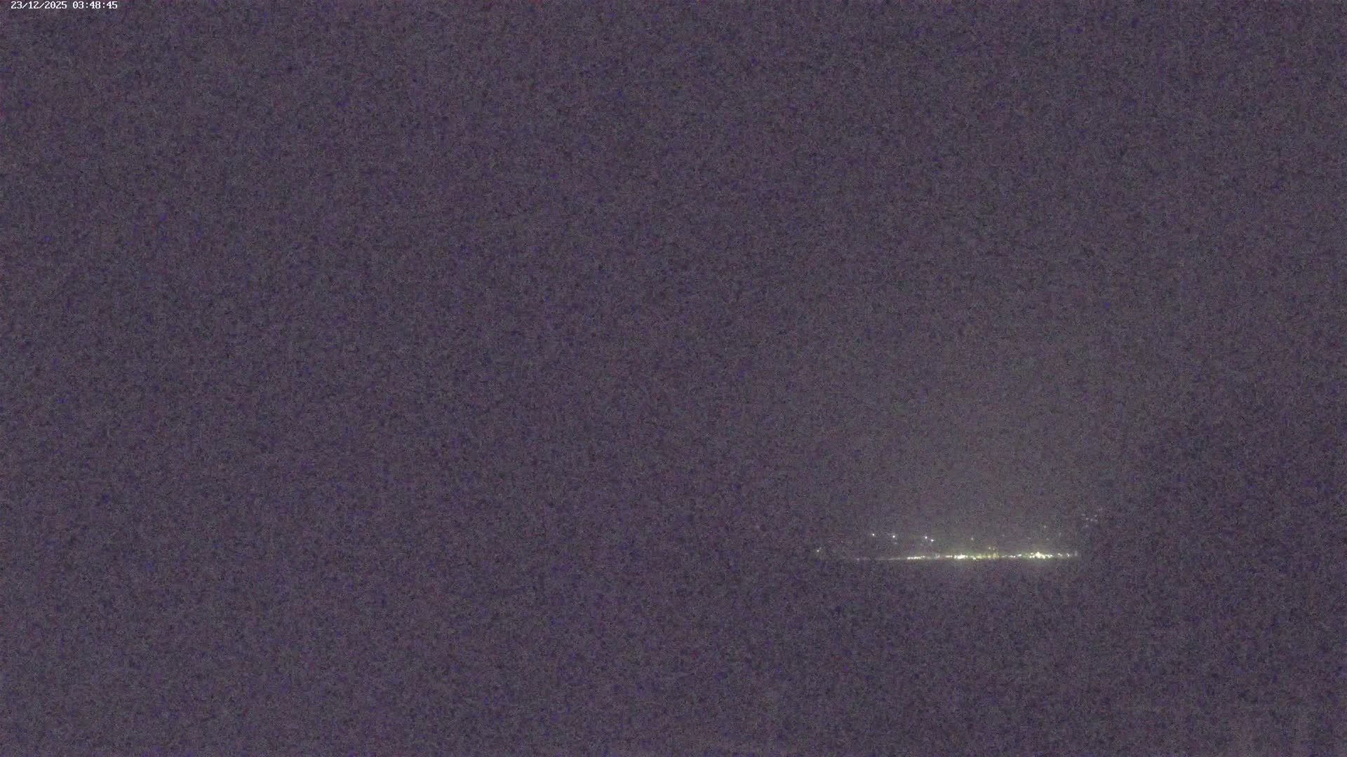 A very dark outdoor night scene reveals a faint horizontal line of distant bright lights against a dark, featureless, and possibly hazy sky.