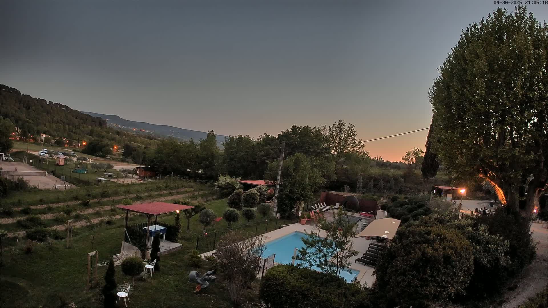An elevated view at dusk shows a swimming pool and surrounding garden area, with hills and trees visible in the background under a clear, twilight sky.