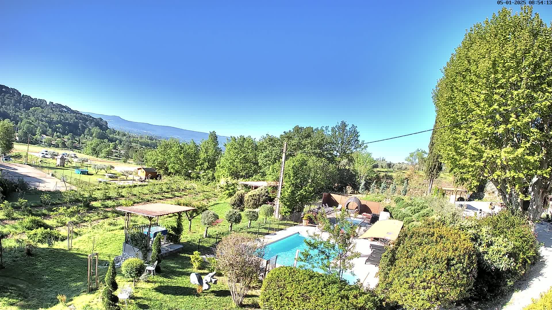 A swimming pool sits in a lush green garden overlooking a hillside under a clear blue sky.