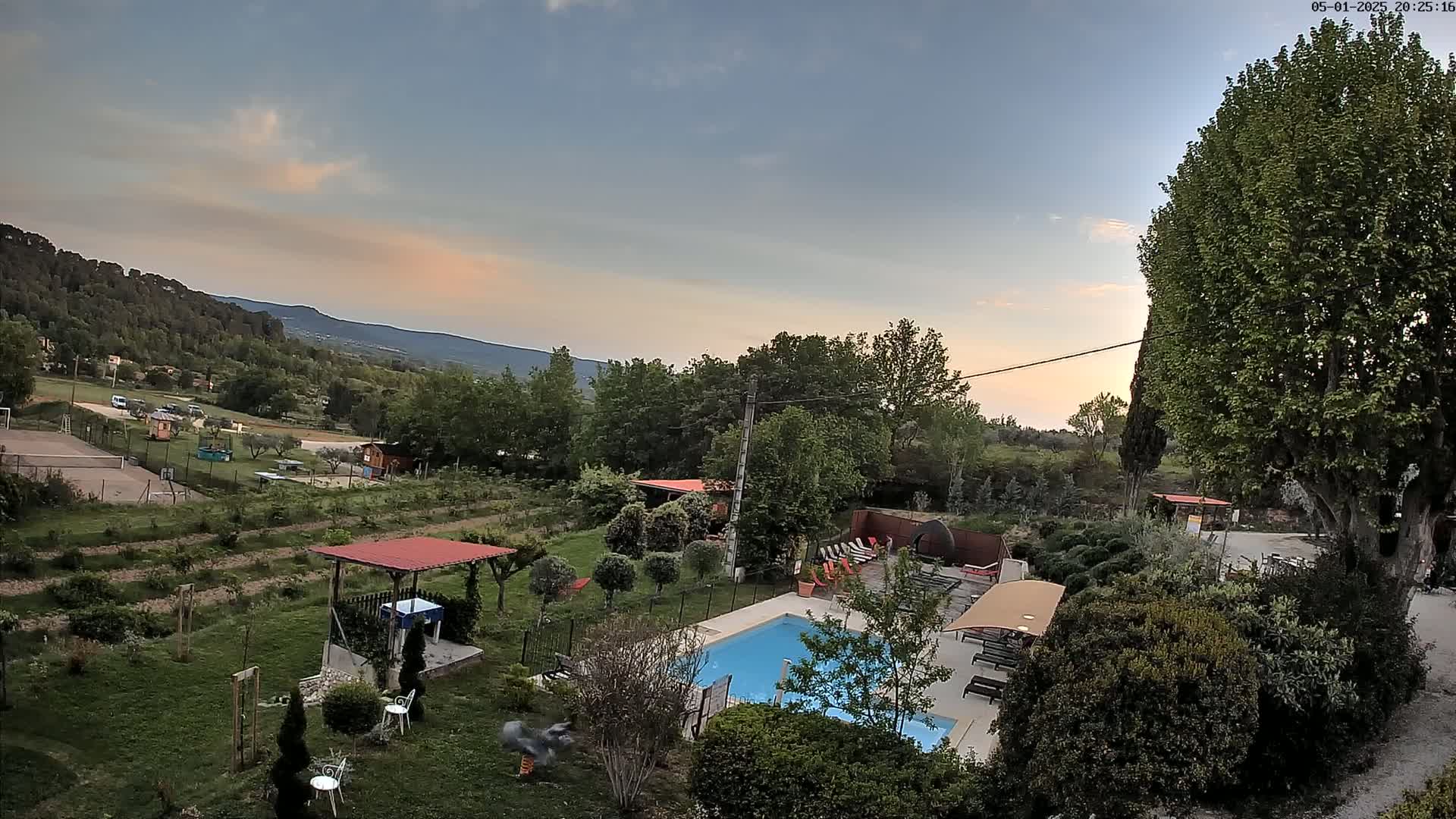 An aerial view of a rural property at sunset shows a swimming pool, lawn, landscaping, and a tennis court, all overlooking a mountain range under a partly cloudy sky.