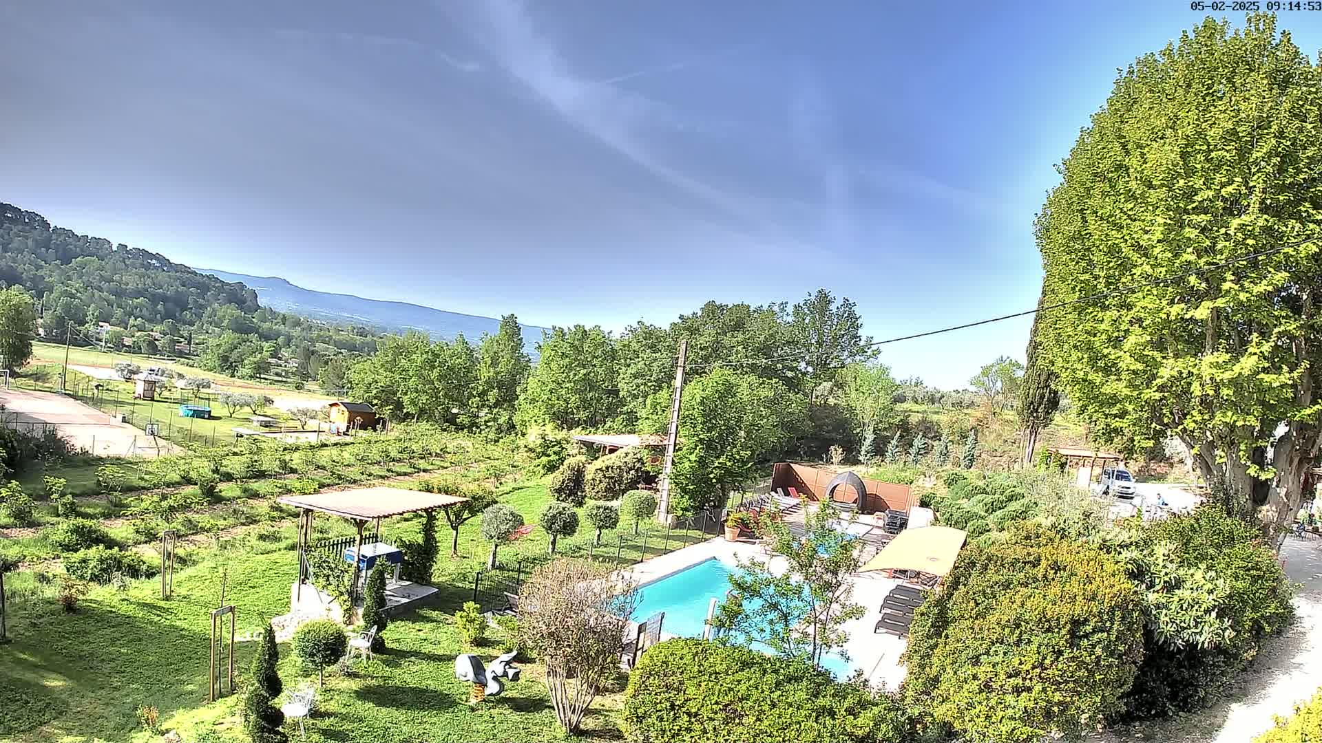 An aerial view shows a swimming pool and surrounding garden nestled in a lush green landscape under a clear blue sky, with distant mountains visible in the background.
