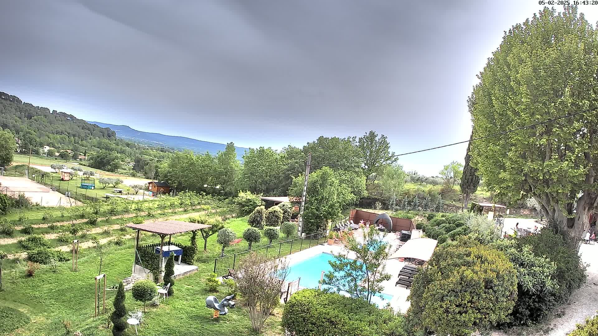 An aerial view shows a swimming pool and surrounding lush greenery under an overcast sky, with distant mountains visible in the background.