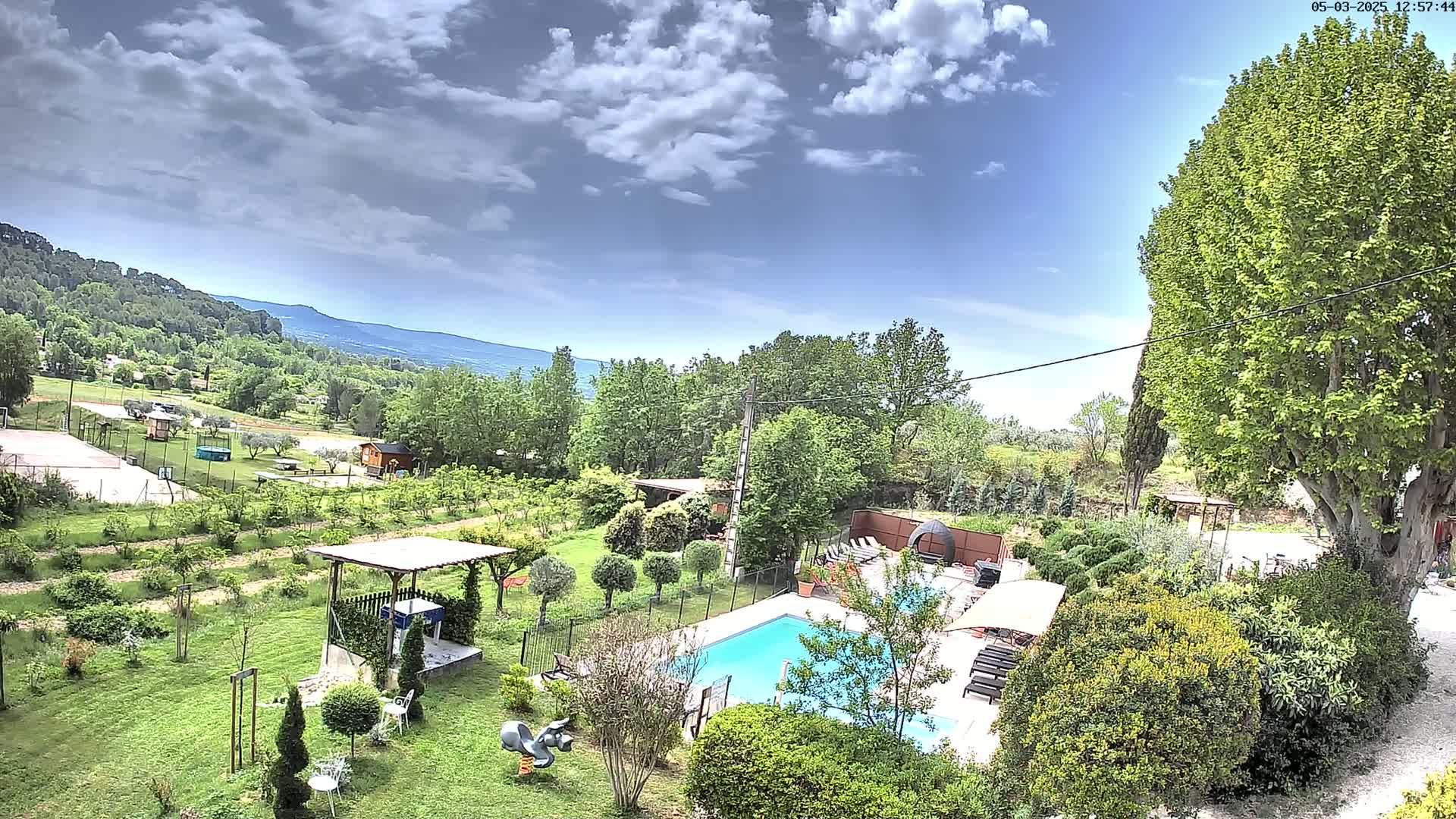 An aerial view shows a swimming pool and lush green landscaping, including a gazebo and playground equipment, with a partly cloudy sky and distant mountains in the background.