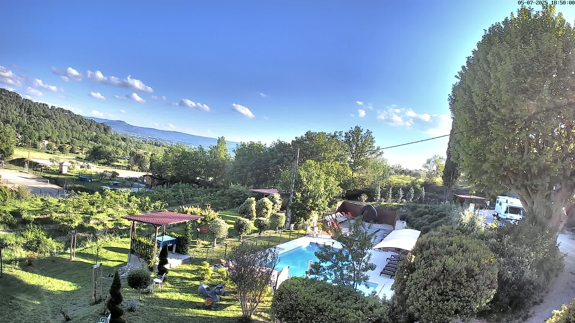 A swimming pool sits in a lush, green garden overlooking a hilly landscape under a mostly sunny sky.