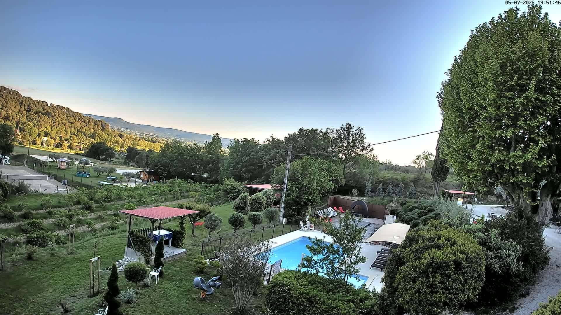 An aerial view of a backyard with a swimming pool, surrounded by lush greenery, overlooking a mountain range under a clear blue sky.