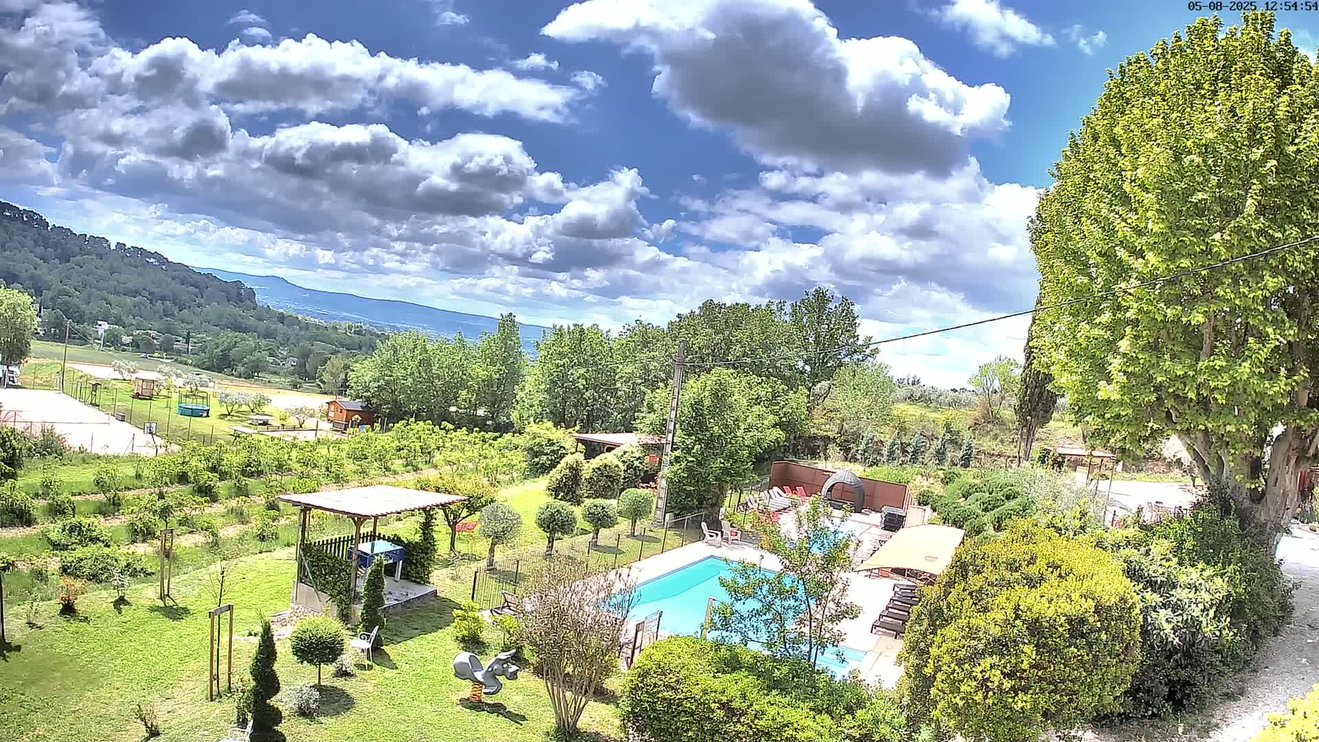 A partly sunny day reveals a swimming pool surrounded by lush greenery, a playground, and a pergola, with distant hills and a partly cloudy sky visible in the background.