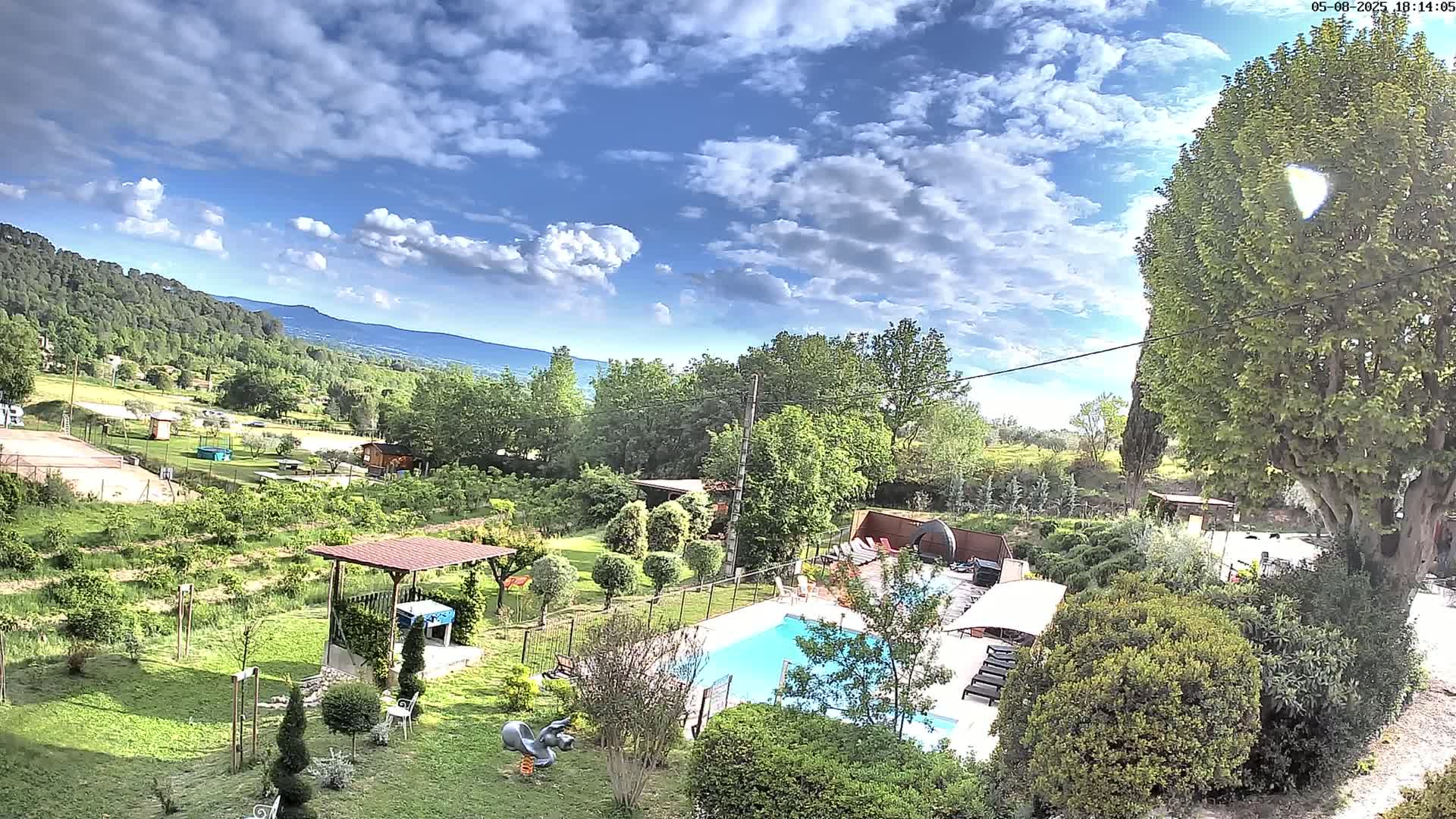 A swimming pool sits in a lush green garden, overlooking a valley under a partly cloudy blue sky.