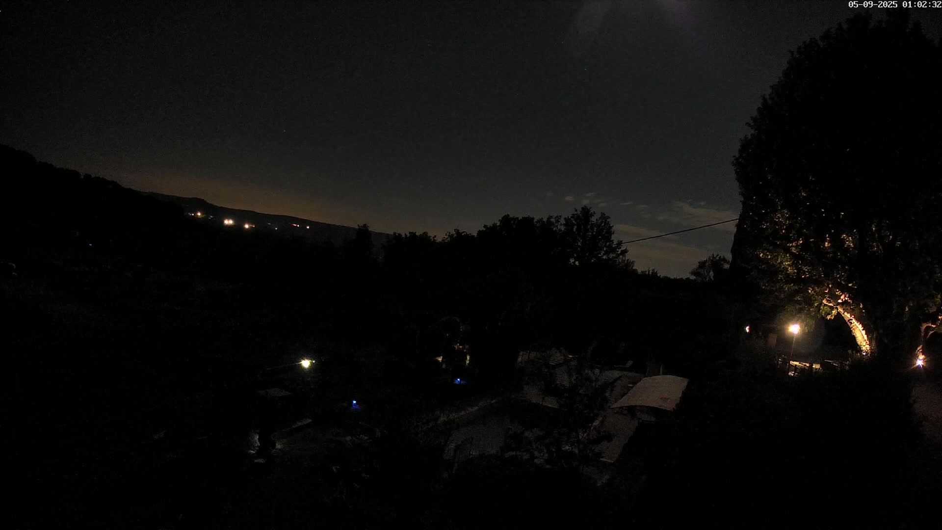 A nighttime view of a dark landscape with trees and dimly lit structures, under a mostly clear sky.