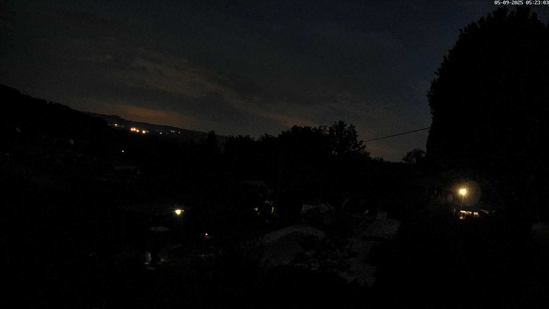 A nighttime view of dark trees and hills, with a few distant lights visible under a mostly clear, dark sky.