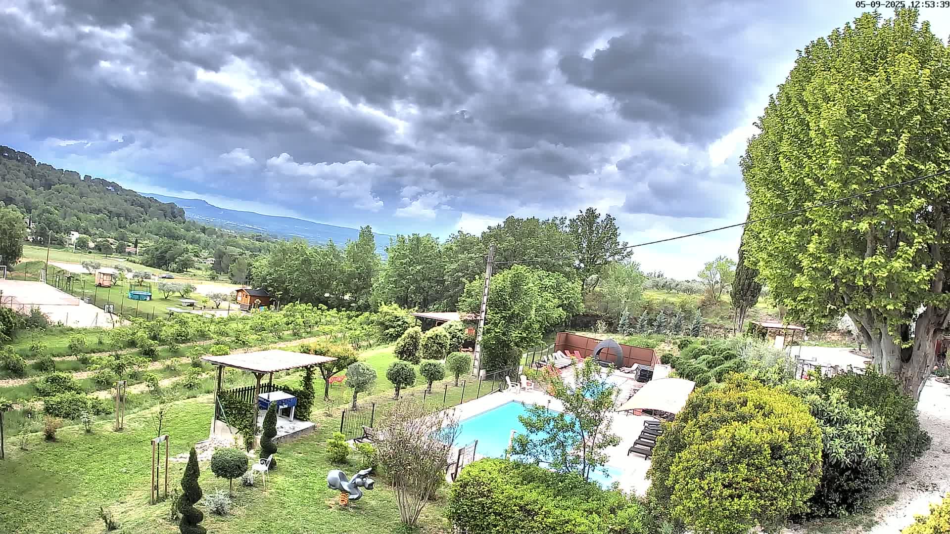 A swimming pool sits in a lush green garden overlooking a hilly landscape under a partly cloudy sky.