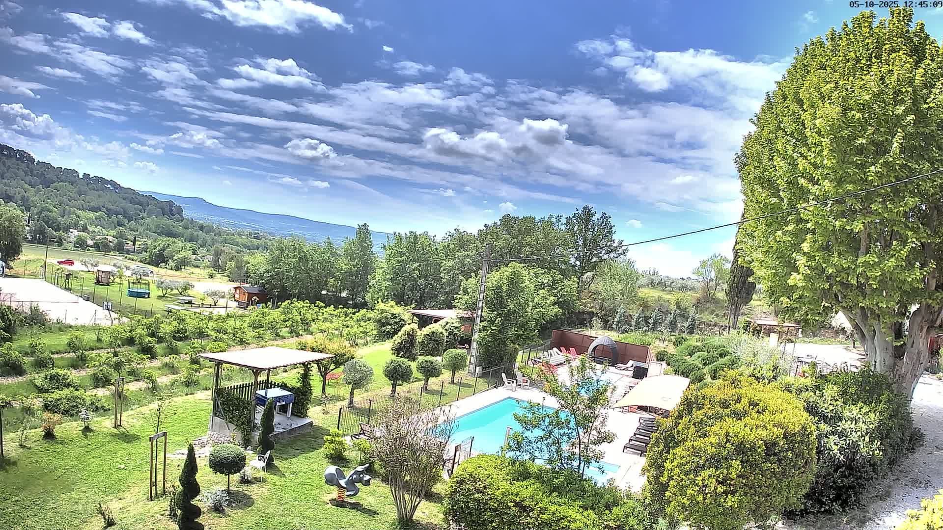A swimming pool and surrounding lush green gardens are nestled in a hillside landscape under a partly cloudy sky.