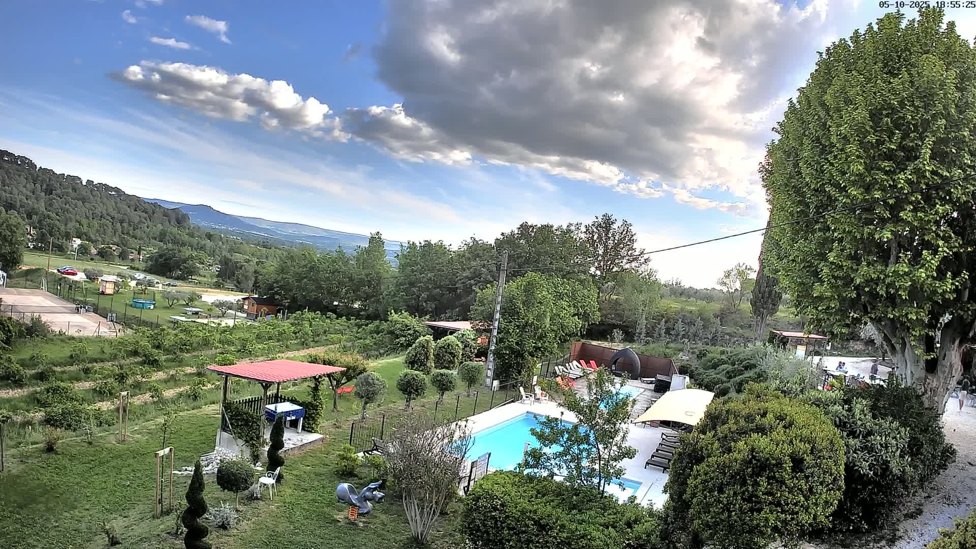 A swimming pool surrounded by a lush, green garden with a view of a partly cloudy sky and distant hills.