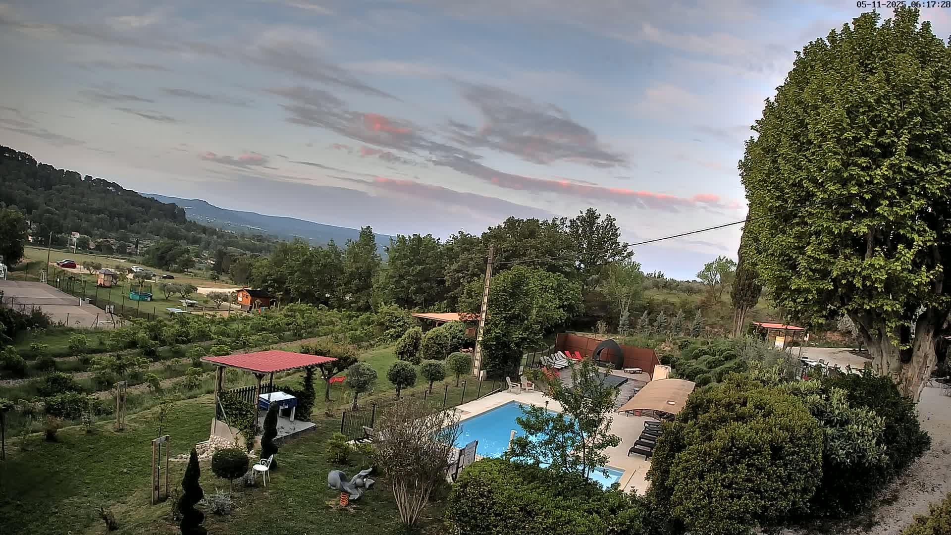 An aerial view of a backyard with a swimming pool, landscaping, and outdoor structures, overlooks a mountainous landscape under a partly cloudy sky with a hint of pink in the clouds.