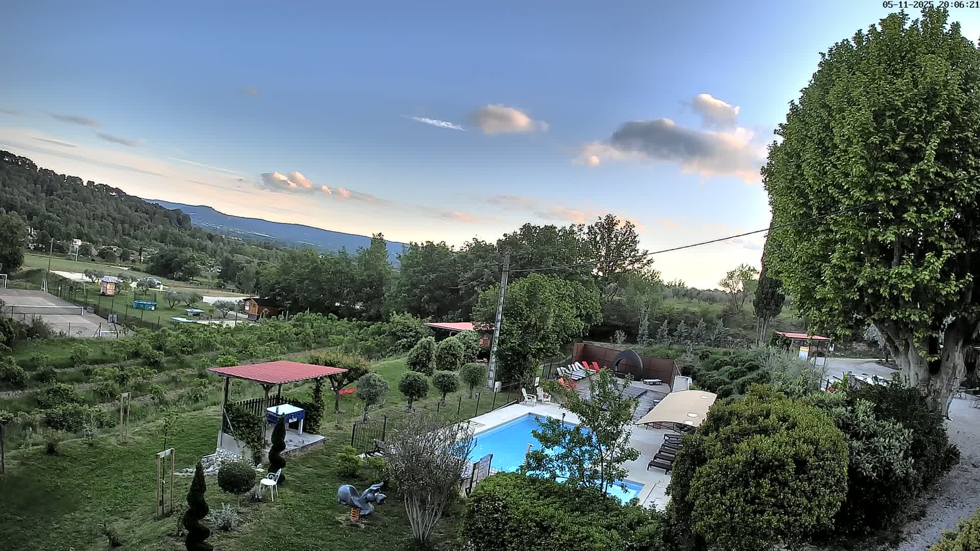 An aerial view shows a swimming pool and surrounding garden, with a tennis court and distant mountains visible under a partly cloudy sky.