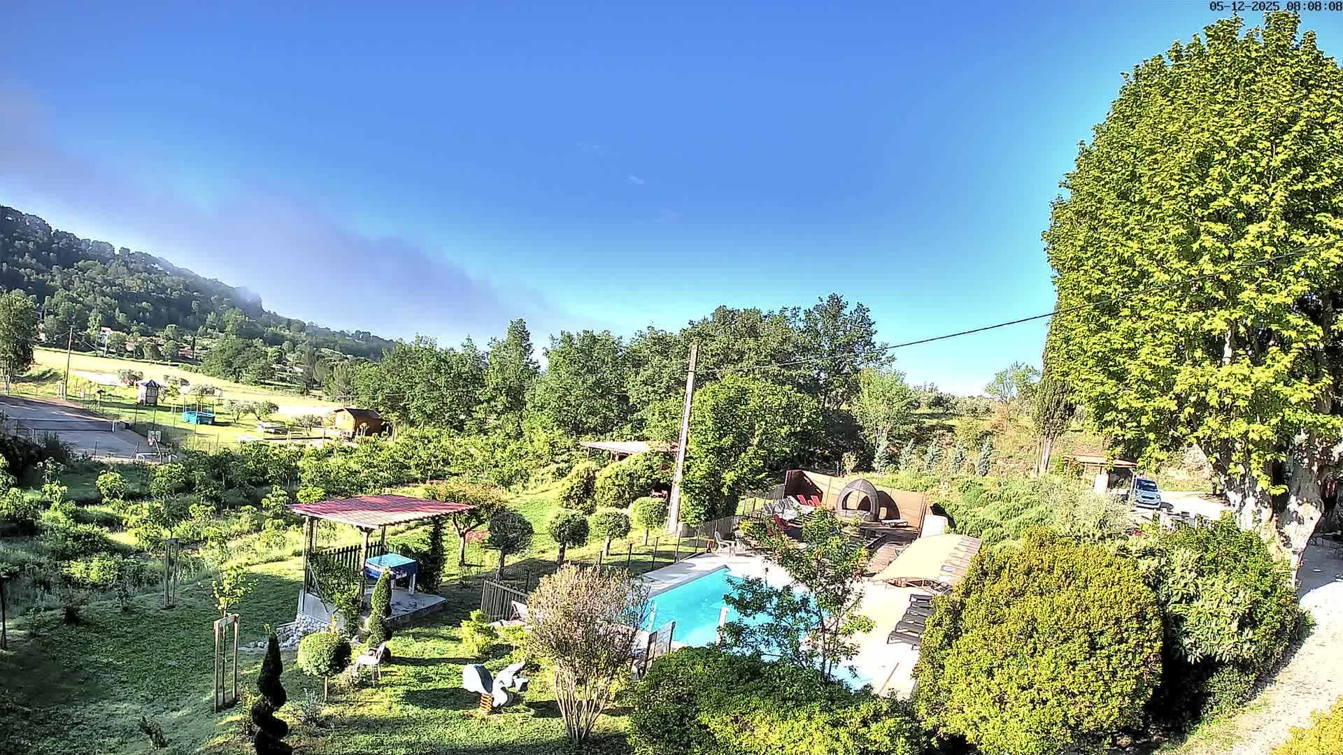 A swimming pool sits in a lush green garden, nestled amongst trees and a hillside under a clear blue sky.