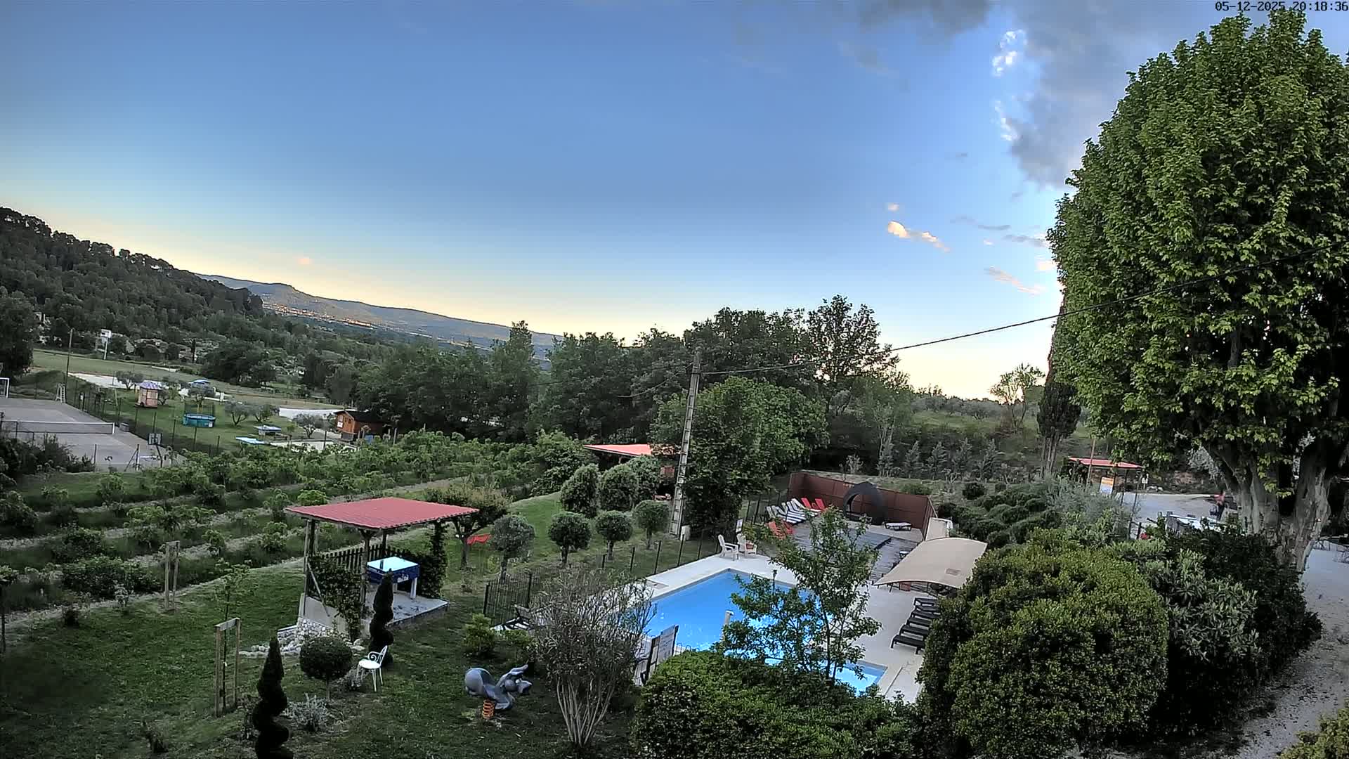 An aerial view shows a swimming pool and surrounding yard with lush greenery and a distant mountain range under a mostly clear evening sky.