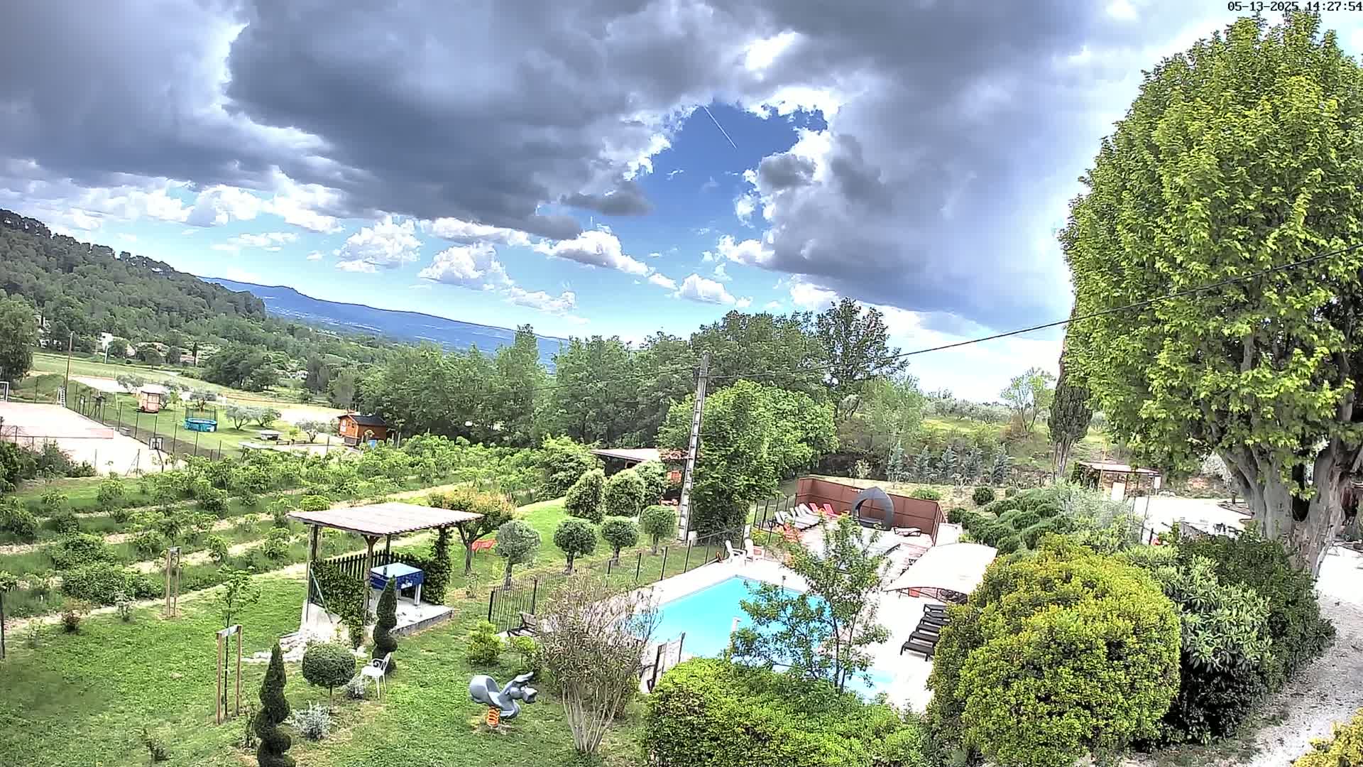 A swimming pool is situated in a lush green garden, overlooking a hilly landscape under a partly cloudy sky.