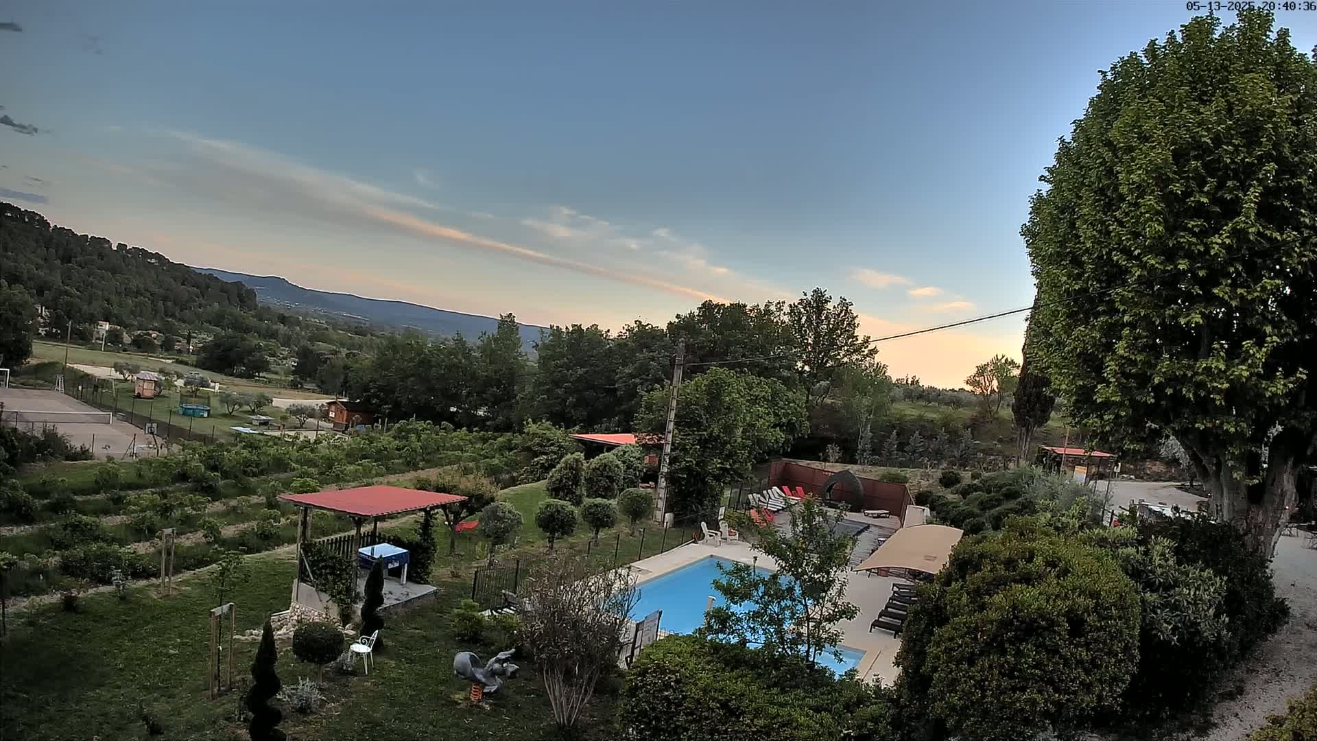 An elevated view of a swimming pool and surrounding garden in a rural setting at sunset, overlooking a valley and mountains under a mostly clear sky.