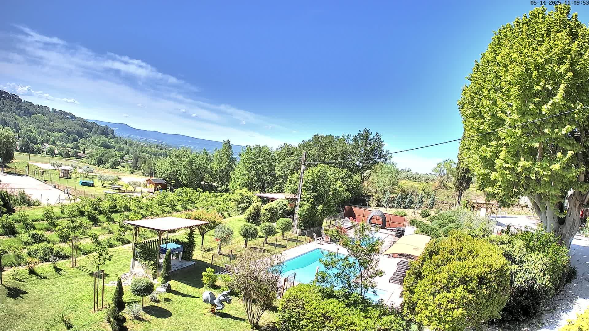 A swimming pool sits in a lush green garden overlooking a valley under a mostly sunny sky.