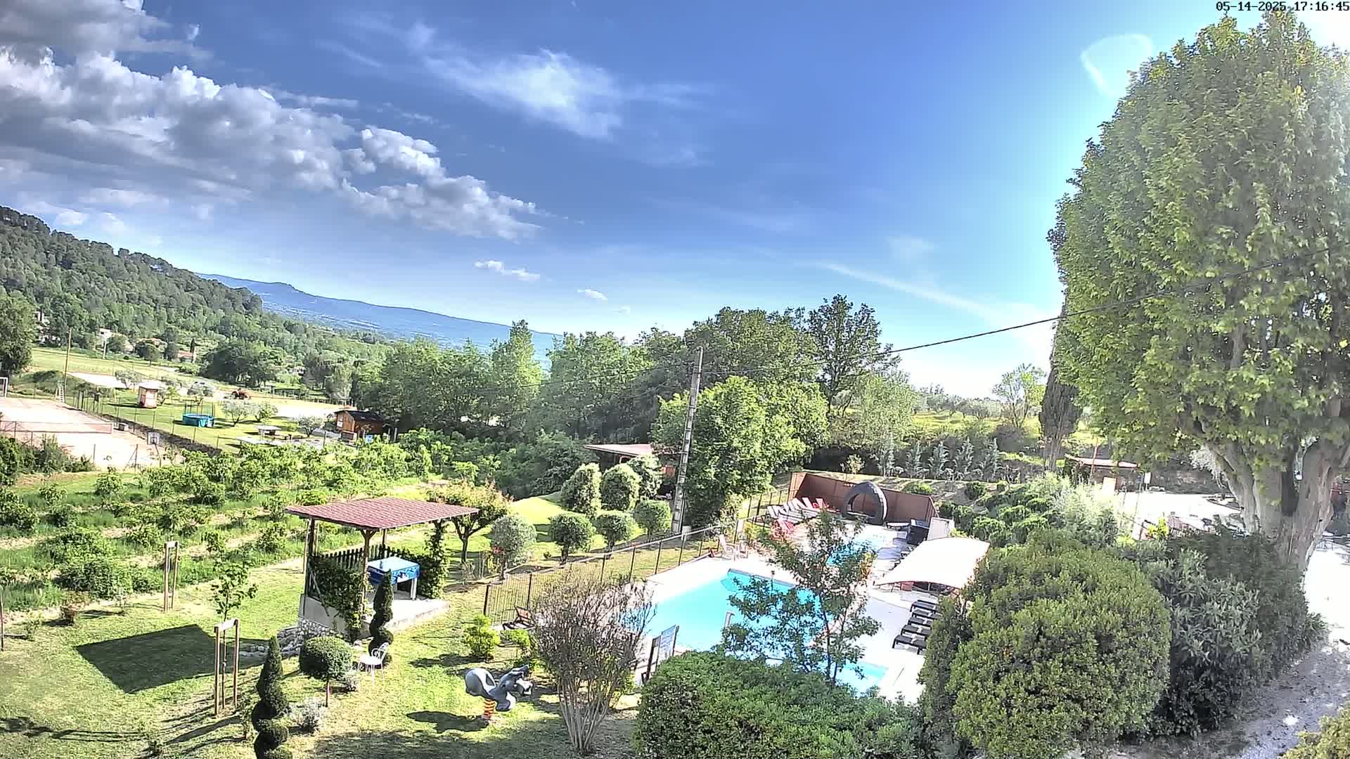 An aerial view shows a swimming pool surrounded by a lush green garden, a pergola, and other yard features, all overlooking a mountainous landscape under a partly sunny sky.