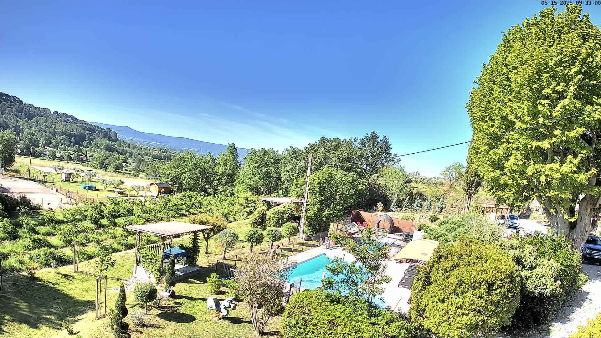 A swimming pool sits in a lush green garden with a pergola, surrounded by trees and shrubs, overlooking a distant mountain range under a clear blue sky.