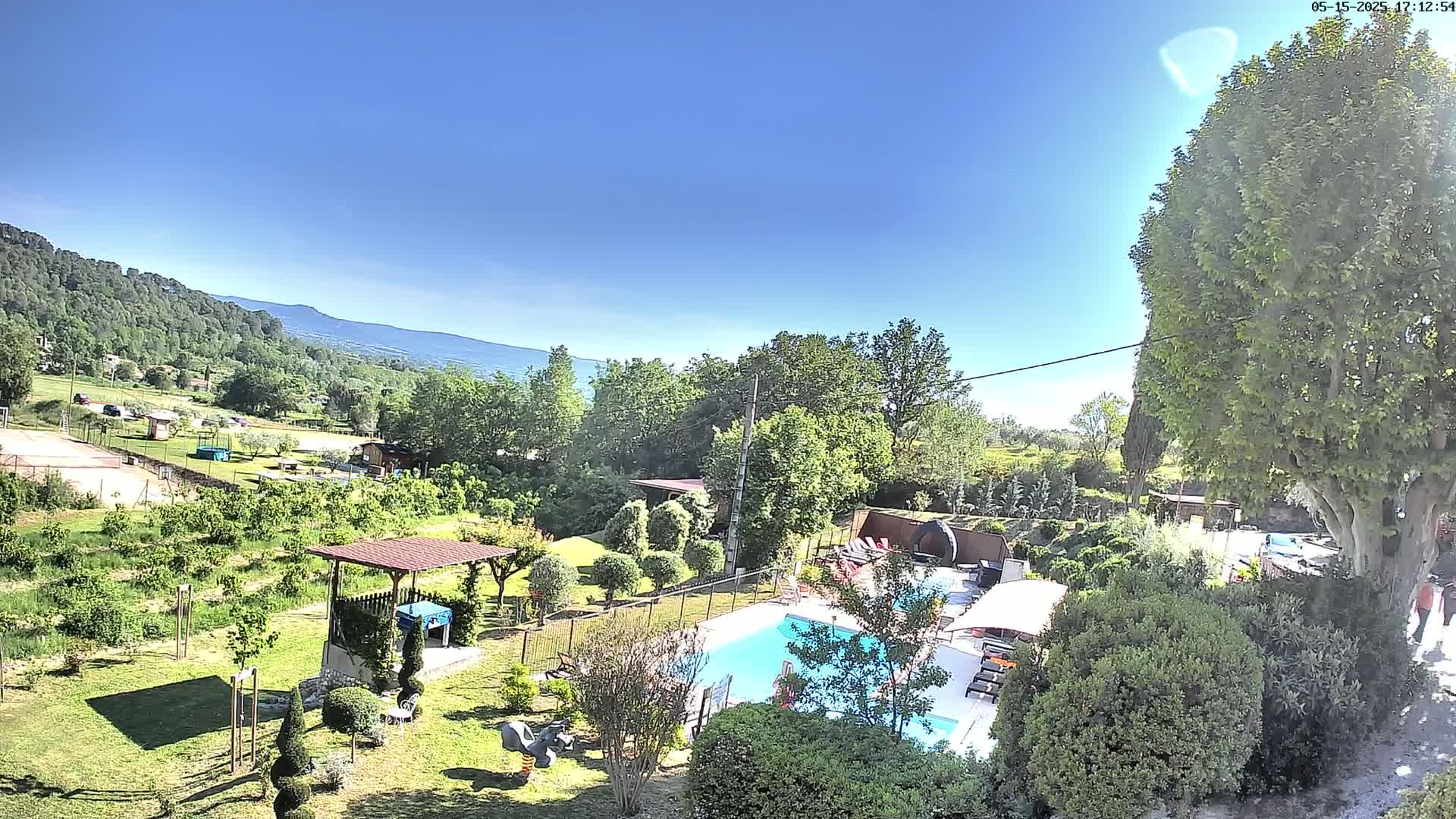 A swimming pool sits in a lush, green garden surrounded by trees and a distant mountain range under a clear, sunny sky.
