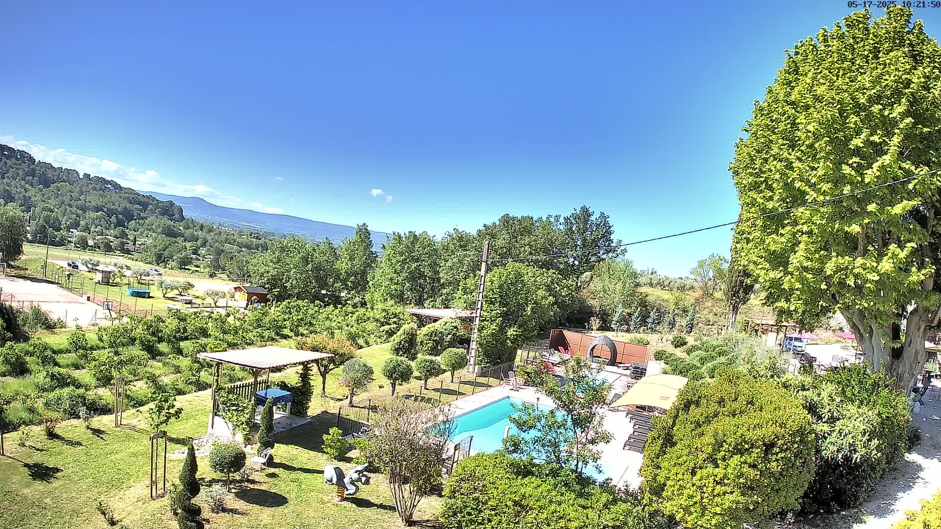 An aerial view shows a swimming pool and surrounding gardens nestled amongst lush greenery and trees under a clear blue sky, with distant mountains visible in the background.