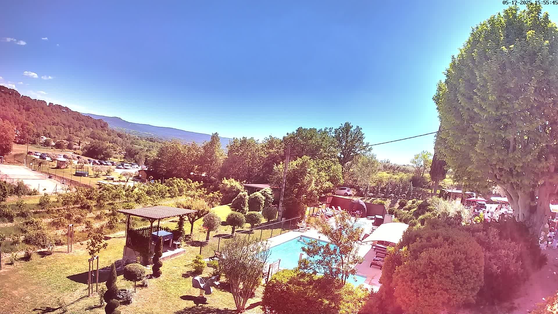 An outdoor scene on a sunny day shows a swimming pool, surrounding gardens, a parking lot, and distant mountains.