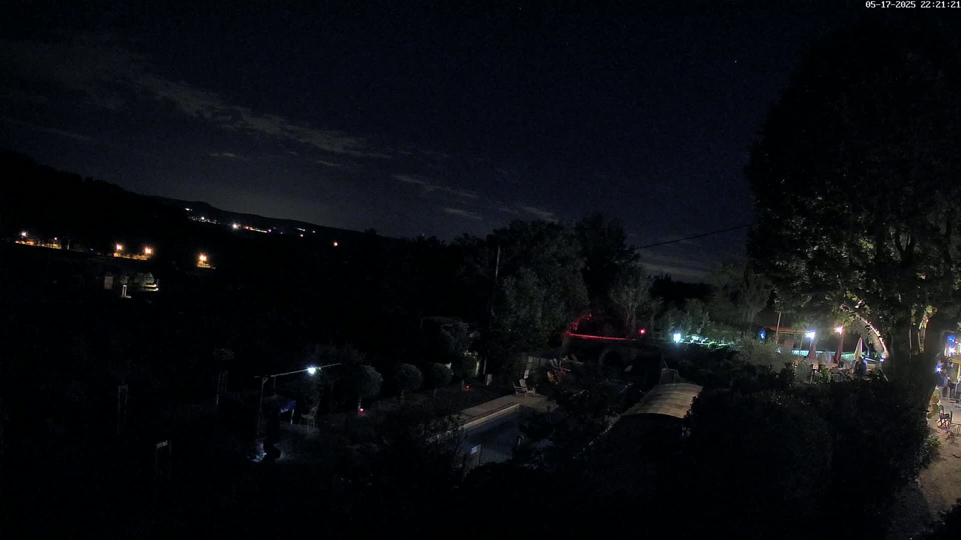 A nighttime view of a backyard with a swimming pool, trees, and some lights, overlooking a dark hillside with distant lights under a mostly clear sky.