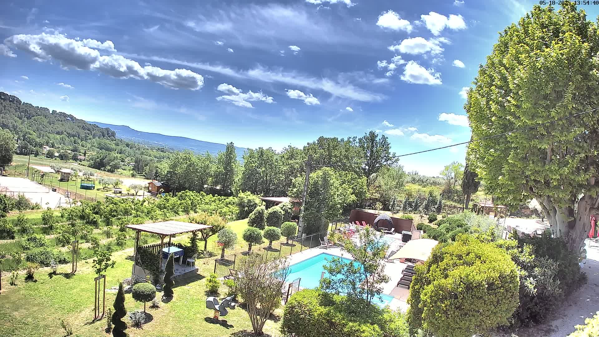 An aerial view shows a swimming pool and surrounding gardens on a sunny day with partly cloudy skies, overlooking a landscape of trees and distant mountains.