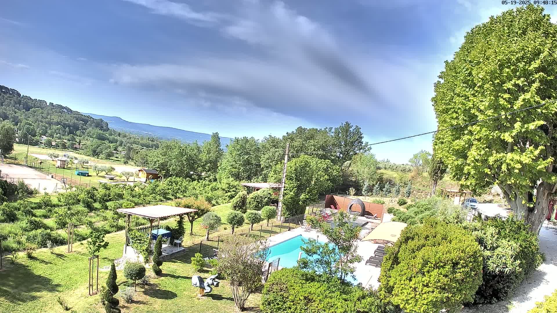 An aerial view shows a swimming pool and landscaped garden with a pergola, nestled amongst lush green trees and hills under a partly cloudy sky.