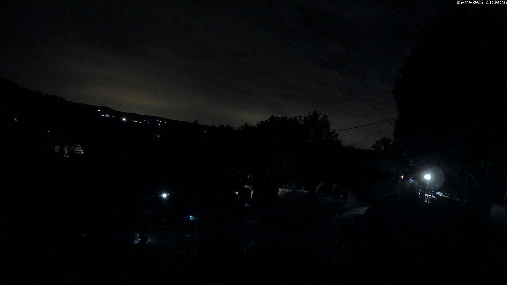 A nighttime view shows a dark landscape with a few distant lights, silhouetted trees, and a single bright light closer to the camera under a mostly cloudy sky.