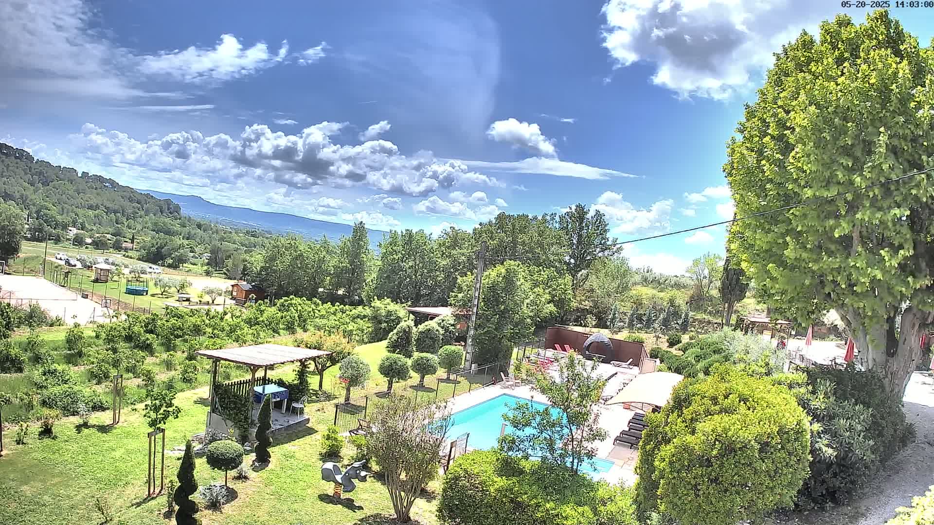 A swimming pool sits in a lush green garden, overlooking a hilly landscape under a partly cloudy, sunny sky.