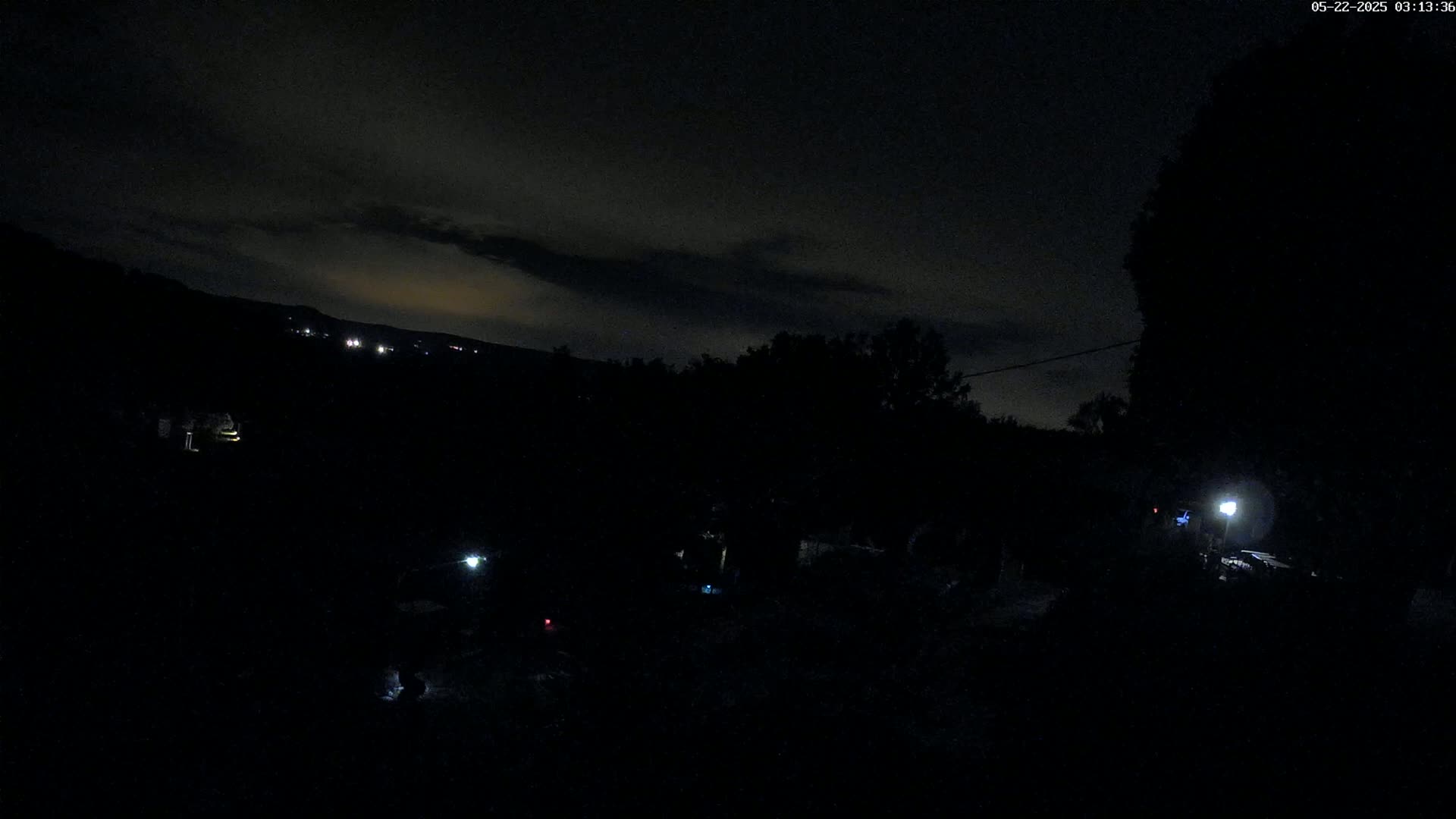 A dark, nighttime scene shows a hilly landscape with sparsely lit areas and a few illuminated objects in the foreground under a mostly cloudy sky.