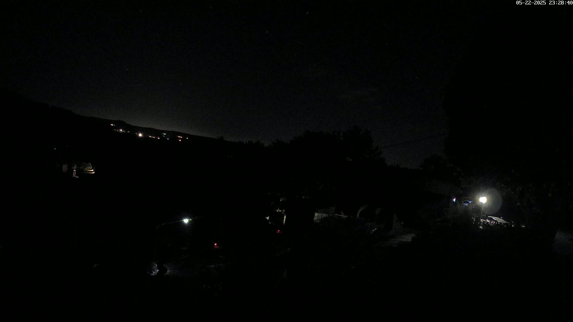A nighttime view shows a dark landscape with a few faint lights in the distance and a brighter light closer to the camera, under a mostly clear, starlit sky.