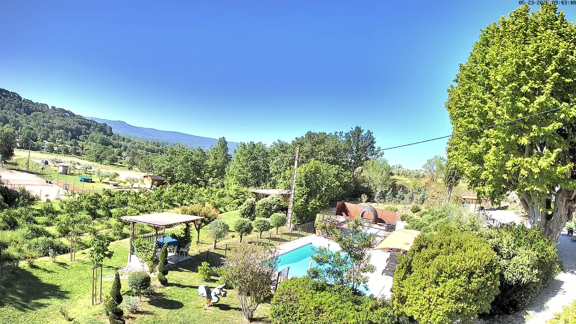 A sunny day reveals a swimming pool nestled in a lush green garden, overlooking a landscape of rolling hills and distant mountains.