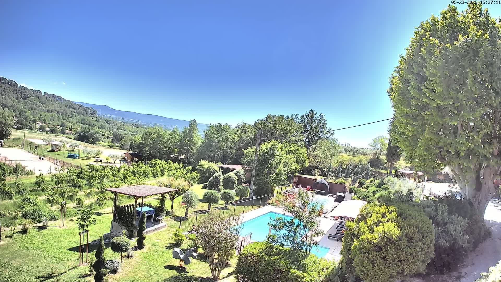 A swimming pool sits in a lush green yard, surrounded by trees and a view of distant mountains under a clear blue sky.