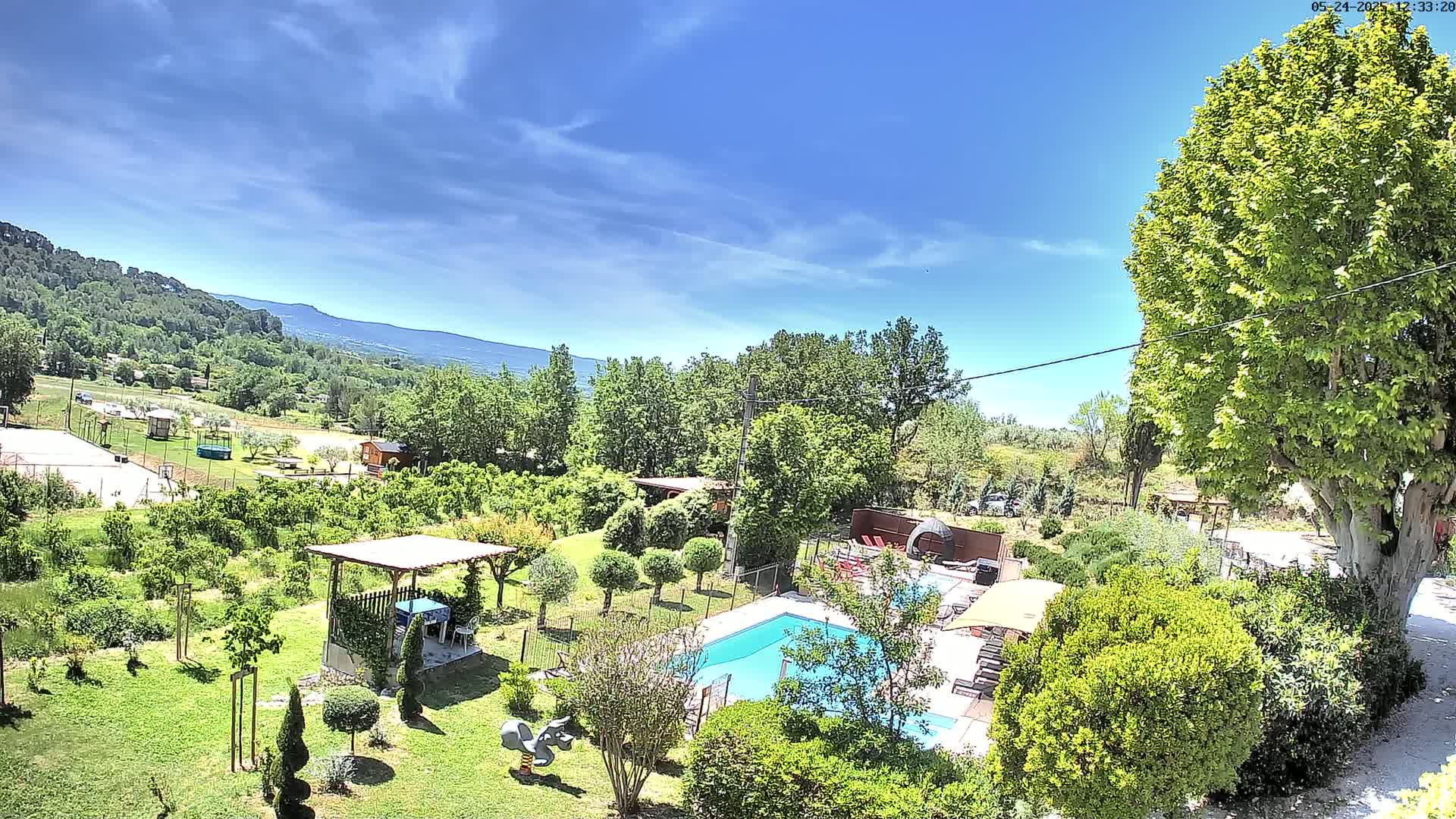 An aerial view shows a swimming pool and surrounding garden on a sunny day, with a verdant landscape and distant mountains visible in the background.