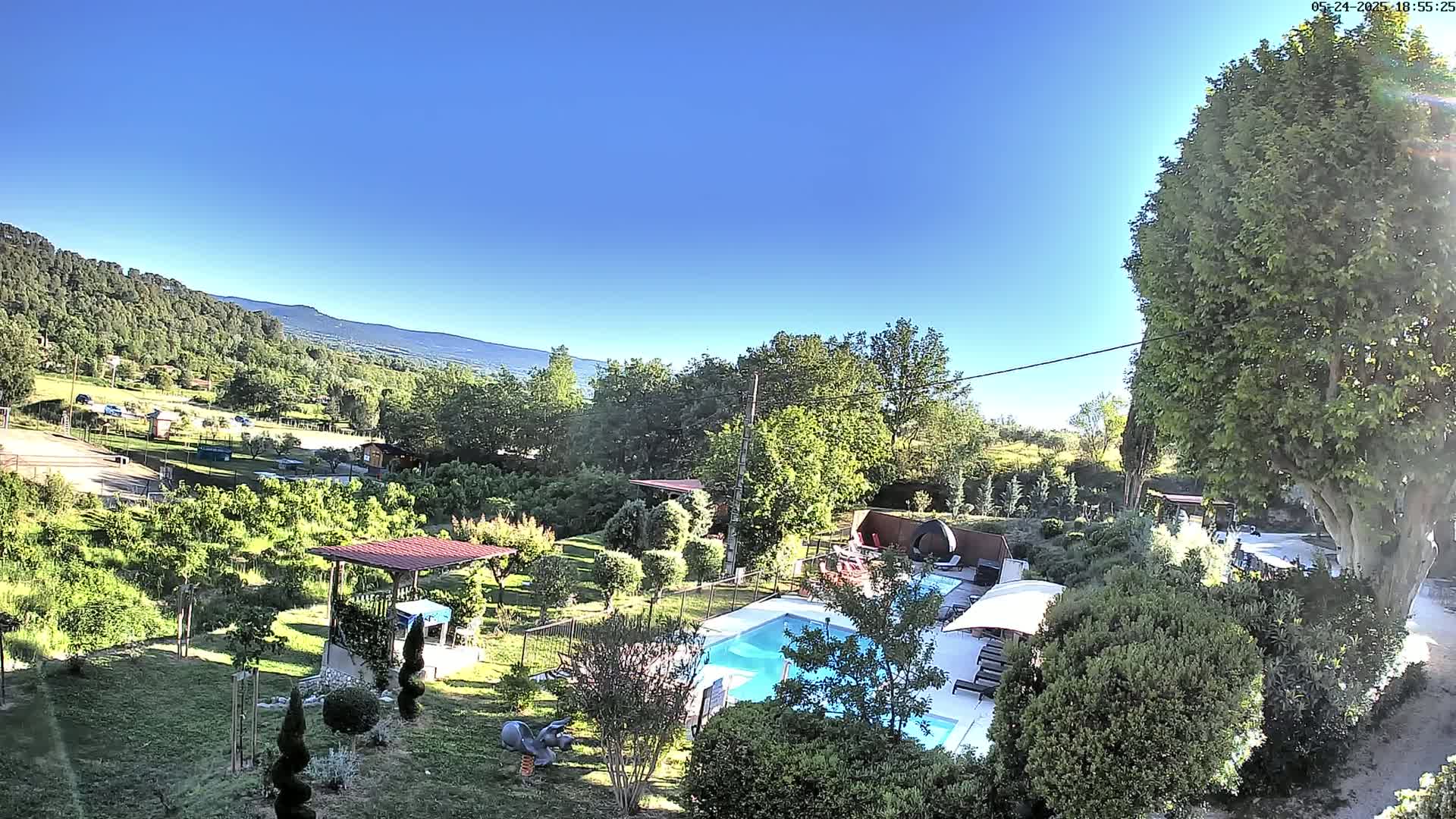 An aerial view shows a swimming pool and surrounding lush gardens, under a clear blue sky, with mountains in the background.